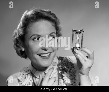 1950s SMILING WOMAN HOLDING UP MINIATURE SIZE THREE MINUTE HOURGLASS EGG TIMER Stock Photo