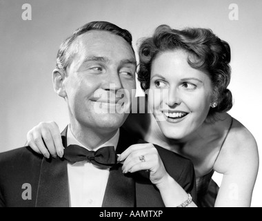 1950s 1960s SMILING COUPLE DRESSED IN FORMAL EVENING ATTIRE WOMAN PULLING ON MAN'S BOW TIE Stock Photo