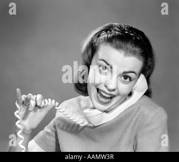 1950s 1960s YOUNG WOMAN TALKING ON TELEPHONE CORD WRAPPED IN FINGERS LOOKING AT CAMERA Stock Photo