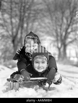 1930s SMILING BOY AND GIRL ON SLED IN SNOW LOOKING AT CAMERA Stock Photo