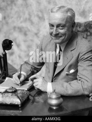 1920s 1930s SMILING MAN BUSINESSMAN SALESMAN SITTING AT DESK SMOKING CIGAR WRITING A MEMO Stock Photo