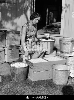 1930s WOMAN FARM WIFE SITTING OUTSIDE OF HEN HOUSE DOORWAY PUTTING EGGS FROM PAILS INTO SHIPPING CRATES Stock Photo