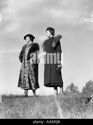 1930s 2 WOMEN POSING OUTSIDE GRASS FASHION PORTRAIT WOMAN HAT HATS FUR COAT LEOPARD STYLE FASHIONS SKY Stock Photo