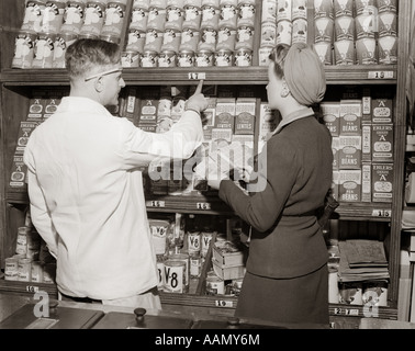 1940s BACK VIEW OF GROCER POINTING OUT PRICES OF CANNED GOODS TO WOMAN HOLDING RATION BOOK Stock Photo