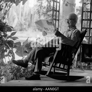 1970s ELDERLY MAN IN ROCKER READING NEWSPAPER ON PORCH Stock Photo