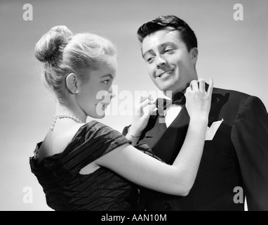 1950s COUPLE IN FORMAL ATTIRE WOMAN WITH BLOND HAIR PULLED BACK IN BUN ADJUSTING MAN'S BOW TIE SMILING PERKY CUTE Stock Photo