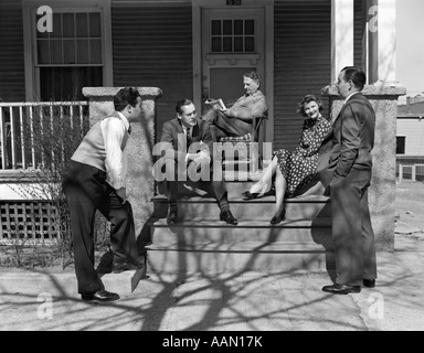 1940s GROUP OF FIVE MEN & WOMEN GATHERED ON & AROUND FRONT PORCH FOUR IN FRONT TALKING ELDERLY MAN IN BACKGROUND READING PAPER Stock Photo