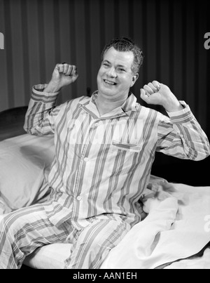 1950s MIDDLE AGED MAN SITTING EDGE OF BED STRIPED PAJAMAS SMILING STRETCHING ARMS BENT SHOULDER Stock Photo