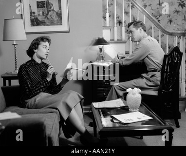 1950s COUPLE IN LIVING ROOM LOOK OVER HOUSEHOLD BILLS FINANCE WOMAN ON COUCH LIST IN HAND MAN SITS AT DESK BY STAIRS Stock Photo