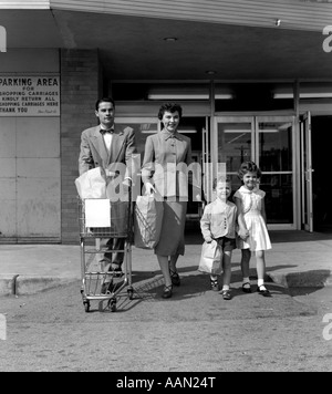 1950s FAMILY MAN WOMAN GIRL BOY WALKING OUT OF SUPERMARKET STORE MOTHER AND SON CARRYING BAGS FATHER PUSHING GROCERY CART Stock Photo
