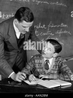 1920s CLASSROOM MALE TEACHER HELPING BOY AT DESK - BLACKBOARD IN BACKGROUND Stock Photo