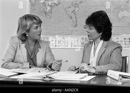 1980s PAIR OF FEMALE TEACHERS HAVING DISCUSSION NOTEBOOKS SPREAD OUT ON DESK IN FRONT OF THEM WORLD MAP ON WALL IN BACKGROUND Stock Photo