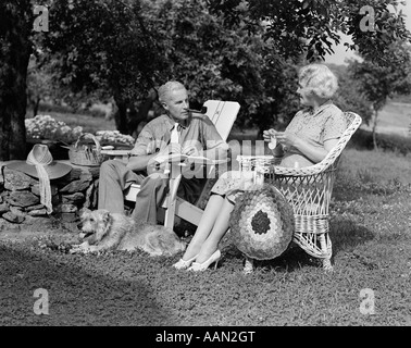 1930s 1940s SENIOR RETIRED COUPLE SITTING OUTDOOR LAWN CHAIRS MAN SMOKING PIPE WOMAN KNITTING WITH DOG Stock Photo