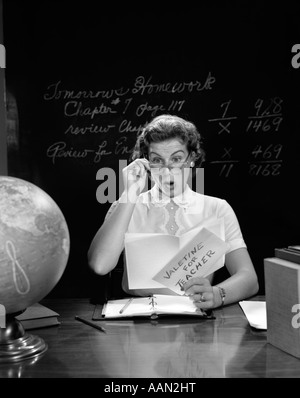 1950s SCHOOL TEACHER AT DESK HAND TO GLASSES EXPRESSION OF SURPRISE OPENING A VALENTINE FOR TEACHER Stock Photo