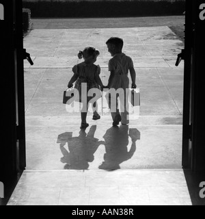 1950s SILHOUETTED BOY GIRL HOLDING HANDS LEAVING SCHOOL DOORS GOING HOME END OF DAY SHADOWS AFTERNOON FRIENDS FRIENDSHIP Stock Photo