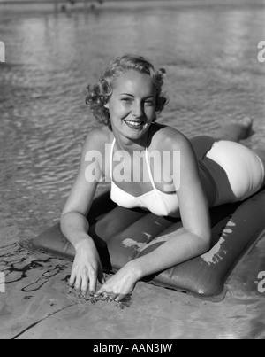 1950s 1940s SMILING BLOND WOMAN WEARING WHITE TWO PIECE BATHING SUIT LAYING ON INFLATED RAFT LOOKING AT CAMERA Stock Photo