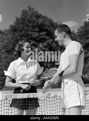 1950s COUPLE STANDING ON OPPOSITE SIDES OF NET HOLDING TENNIS RACQUETS Stock Photo