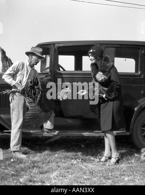1920s FARMER HOLDING TURKEY TALKING TO WOMAN FASHIONABLY DRESSED HAND HELD OUT TO TURKEY IN FRONT OF CAR Stock Photo