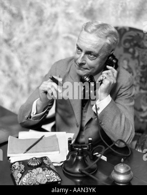 1930s ELDER MAN SITTING AT DESK SMOKING CIGAR TALKING ON TELEPHONE Stock Photo