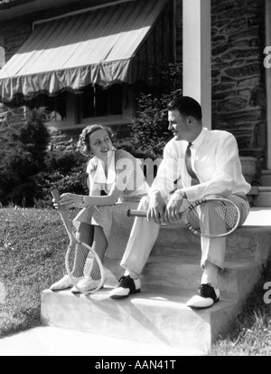 1930s COUPLE HOLDING TENNIS RACKETS WOMAN IN DRESS MAN IN TENNIS WHITES AND SADDLE SHOES SITTING ON STEPS OF HOUSE Stock Photo