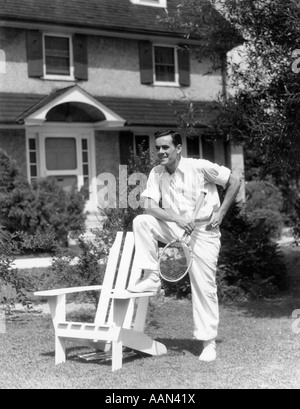 1930s 1940s MAN IN TENNIS WHITES HOLDING RACKET WITH FOOT ON WHITE ADIRONDACK LAWN CHAIR Stock Photo