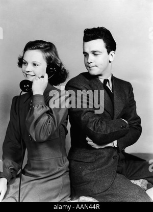 1940s YOUNG COUPLE GIRL TALKING ON TELEPHONE BOY ARMS CROSSED LOOKS ANNOYED Stock Photo