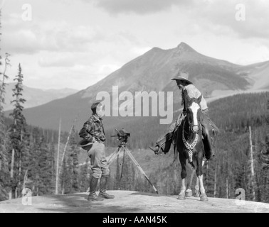 1930s CAMERAMAN IN BOOTS JODHPURS & WOOL HUNTING SHIRT STANDING NEXT TO MOVIE CAMERA ON TRIPOD TALKING TO MOUNTED COWBOY Stock Photo