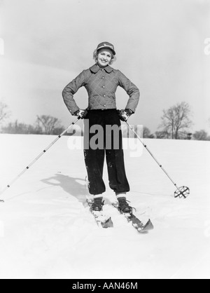 1940s 1950s SMILING WOMAN LOOKING AT CAMERA ON SKIS WITH CAP AND QUILTED SKI JACKET WITH BOTH HANDS ON HIPS Stock Photo