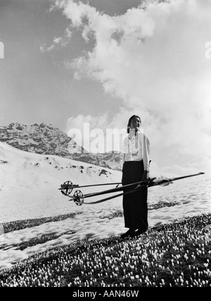 1930s WOMAN HOLDING SKIS POLES STANDING ON CROCUSES IN SNOWY ALPS NEAR DAVOS SWITZERLAND SPRING CHANGE OF SEASONS Stock Photo