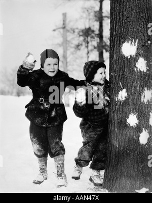 1920s 1930s SMILING BOY AND GIRL PLAYING IN THE SNOW HIDING BEHIND TREE WITH SNOWBALLS IN HAND Stock Photo