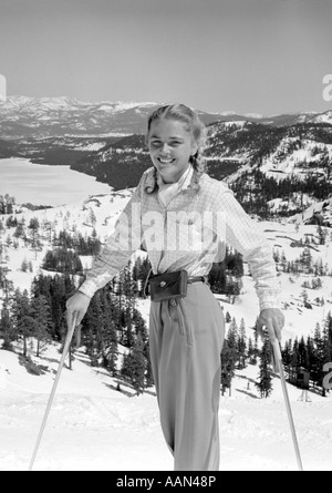 1940s 1950s YOUNG BLOND ATHLETIC WOMAN LOOKING AT CAMERA SMILING STANDING WITH SKI POLES TOP OF MOUNTAIN Stock Photo