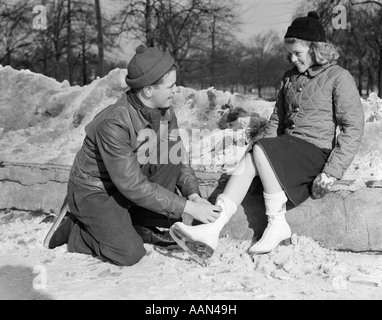 1930s 1940s TEENAGE COUPLE IN KNIT CAPS & WINTER JACKETS KNEELING BOY TYING ICE SKATES FOR GIRL Stock Photo