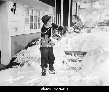 1950s GIRL IN CAP SCARF SWEATER MITTENS & WINTER CLOTHING SHOVELING SNOW OFF STEPS & WALKWAY OF SUBURBAN HOME Stock Photo
