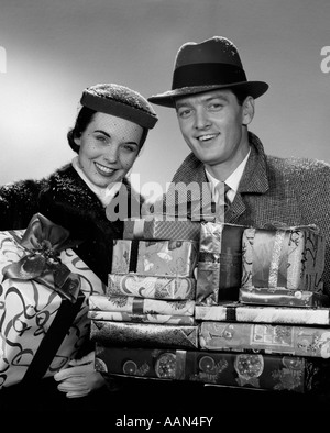 1950s 1960s SMILING PORTRAIT COUPLE IN WINTER COATS & HATS HOLDING WRAPPED CHRISTMAS GIFTS LOOKING AT CAMERA Stock Photo
