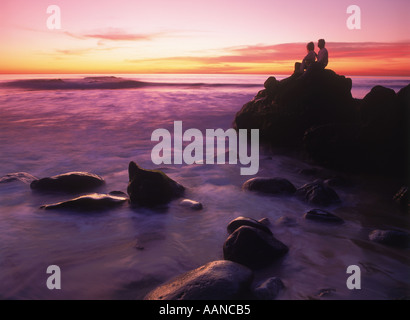 Couple sitting on rocky shore above waves at sunset in California Stock Photo