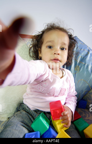 Young baby toddler aged sat on a chair with learning blocks reaching her hand out to grab the camera or viewer