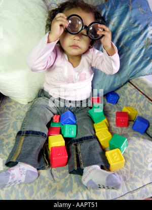 Young baby toddler aged sat on a chair with learning blocks taking thick set glasses off
