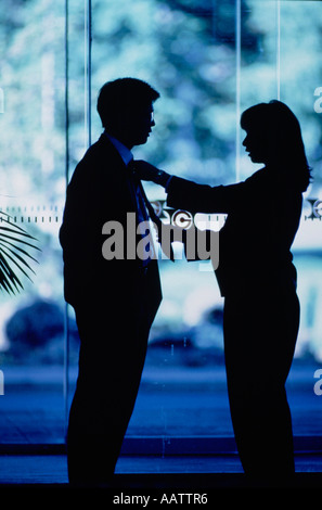 Silhouette profile of woman adjusting man's tie whilst standing in lobby of modern glass-walled building. Stock Photo