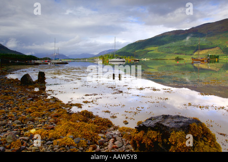 Boats on Loch Leven at Balluchullish with rocky shoreline in foreground and perfect reflections in surface of water Stock Photo