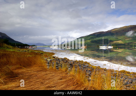 Boats on Loch Leven at Balluchullish with rocky grassy shoreline in foreground and perfect reflections in surface of water Stock Photo