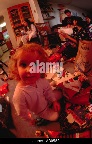 A young girl sits on the floor amidst the torn wrapping paper on Christmas morning as adults sit in the background Stock Photo