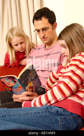 Father reading book to his daughters on sofa in living room at home UK Stock Photo