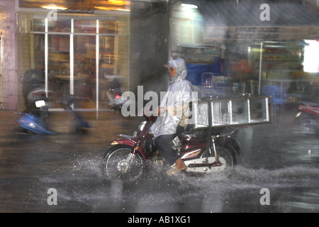 Man in plastic raincoat with motorbike load rides along flooded De Tham Street Pham Ngu Lao district Ho Chi Minh City Vietnam Stock Photo