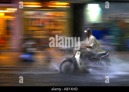 Man in cape on motorcycle rides rain along flooded De Tham Street Pham Ngu Lao district Ho Chi Minh City Vietnam Stock Photo