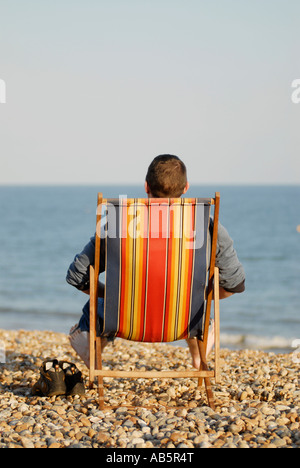 man in deck chair sitting on pebbled beach by the sea Stock Photo