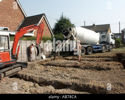 Work in progress delivery truck concrete pouring into trench fill foundation for new detached house with mini excavator on building site plot Essex UK Stock Photo