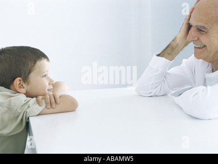 Boy and grandfather sitting across table, smiling at each other Stock Photo