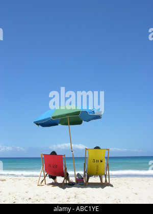 A man and a woman sit on a red and a yellow canvas chair, in the center a sun screen, at the otherwise deserted beach at the Stock Photo