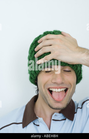 Young man wearing knit hat, hand on head, eyes closed, sticking out tongue, portrait Stock Photo