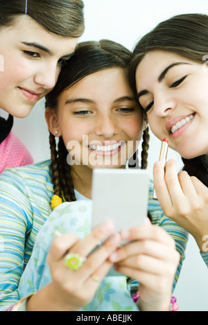 Three young female friends looking at selves in hand mirror, one holding lipgloss Stock Photo